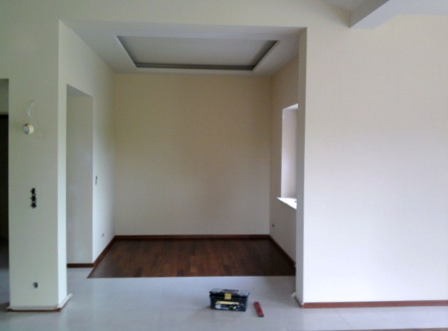 Pomalowane na kremowo wnętrze z drewnianą podłogą, widoczne narzędzia w skrzynce i poziomica, sufit z dekoracyjnym wgłębieniem.